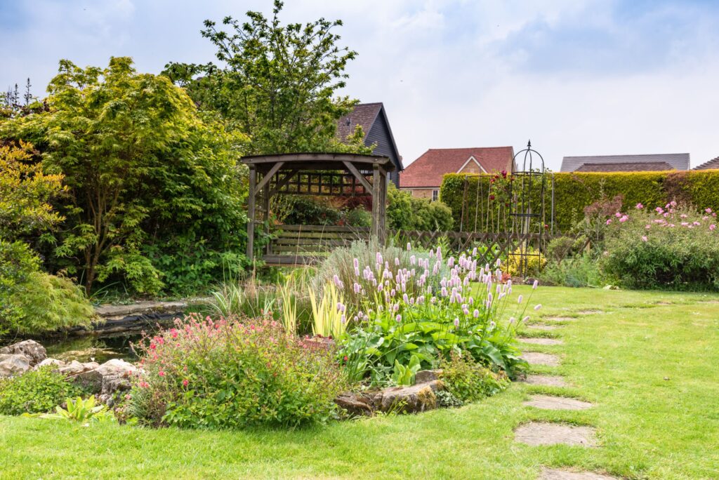 Ein grüner Garten mit Pflanzen, einem kleinen Teich und eine Art Gartenhaus.