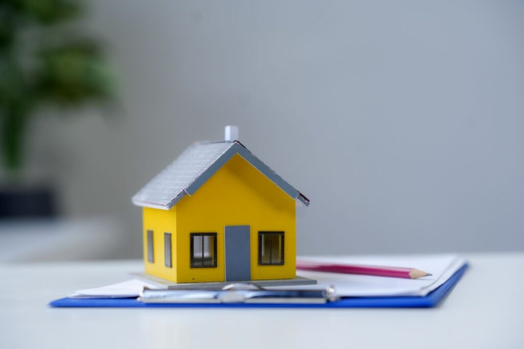 Ein gelbes Miniaturhaus steht auf einem Dokument mit einem rosa Stift und deutet auf Immobilientransaktionen und den Kaufprozess von Eigenheimen hin