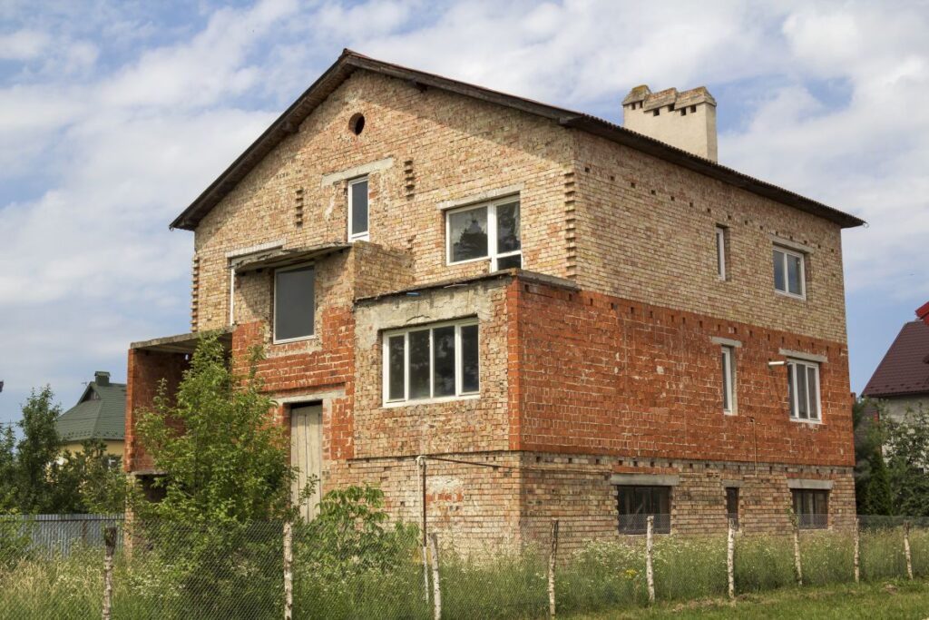 Neues unfertiges Backsteinhaus mit vergittertem Türeingang, Kunststofffenstern im ersten Stock, Ziegel- und Kieshaufen auf grünem Gras auf eingezäuntem Grundstück.