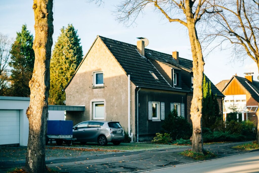 Straße und Einfamilienhaus mit altem Baumbestand und einem Auto in der Auffahrt 