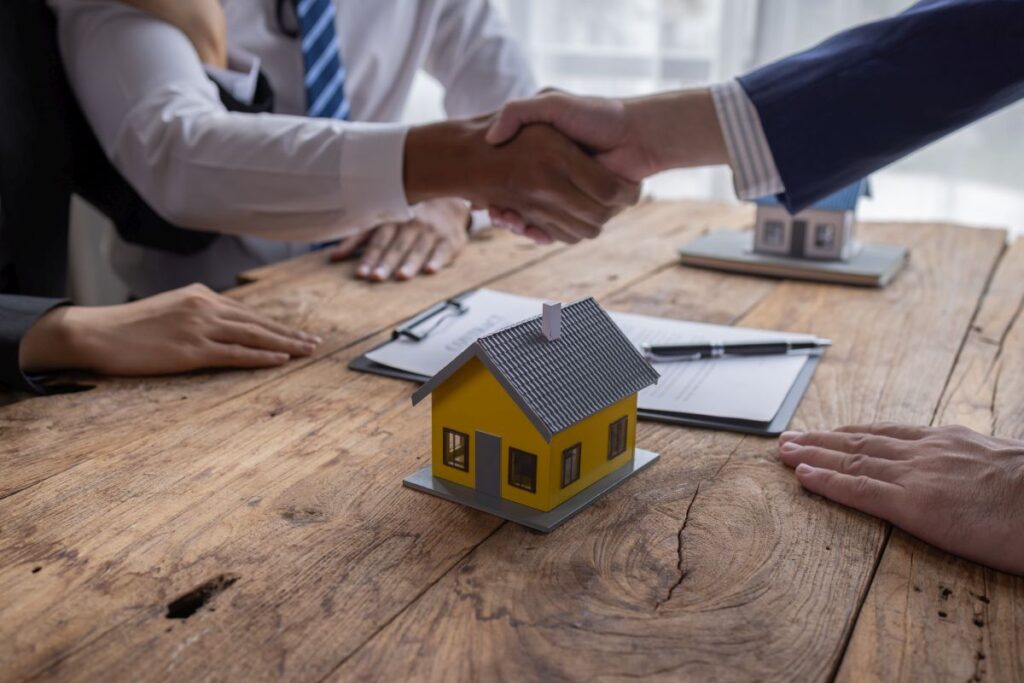 Auf dem Bild schütteln sich zwei Menschen die Hand, auf dem Tisch steht ein kleines Haus und daneben liegt ein Vertrag. 