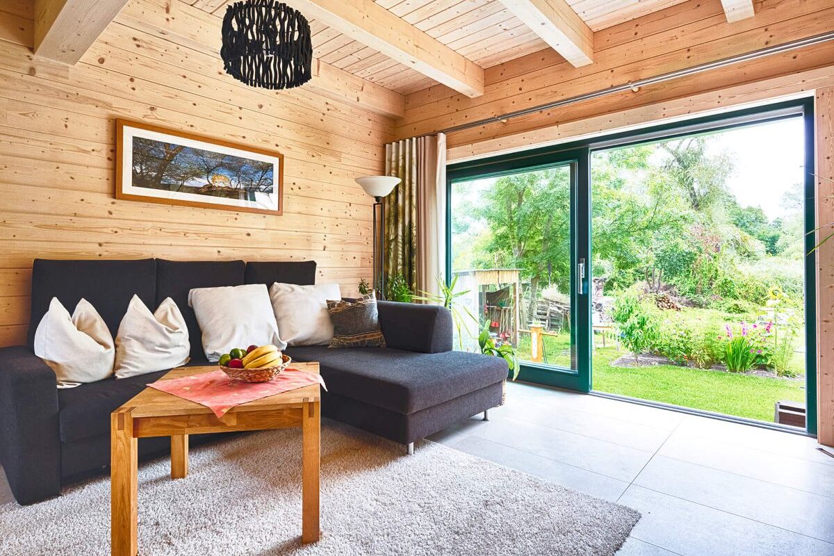Blockhaus Alpina One Wohnzimmer mit Holzdecke und dunklem Sofa und Blick ins Grüne