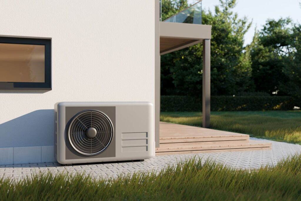 Eine moderne Wärmepumpe steht auf der Terrasse eines Wohnhauses in einem üppig grünen Garten. Das hellgraue Außengerät befindet sich an der Hauswand. Neben dem Gerät ist ein kleines Fenster zu sehen. Im Hintergrund erstrecken sich eine Holzterrasse und eine Grünfläche mit Bäumen. 